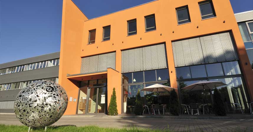 El edificio principal de Munich Enterprise Software con una llamativa fachada naranja y grandes ventanales. En primer plano hay una escultura metálica decorativa, y la zona exterior está equipada con mesas y sombrillas, creando un ambiente acogedor.
